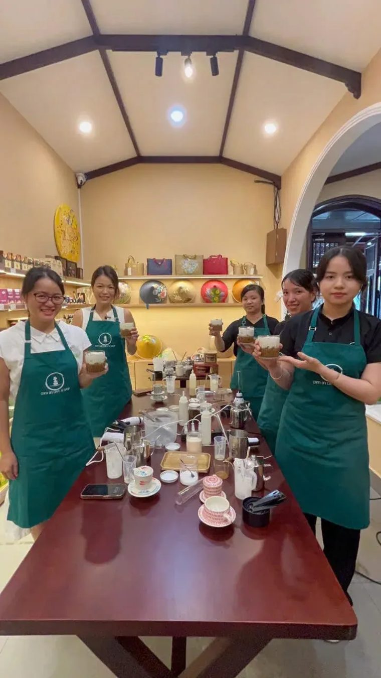 Học viên mặc tạp dề xanh cầm ly cà phê quanh bàn học tại Cam On Cafe.
