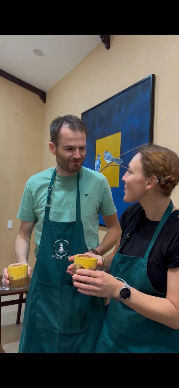 Hai du khách mặc tạp dề xanh cầm ly cà phê trứng vừa pha xong tại Cam On Cafe.