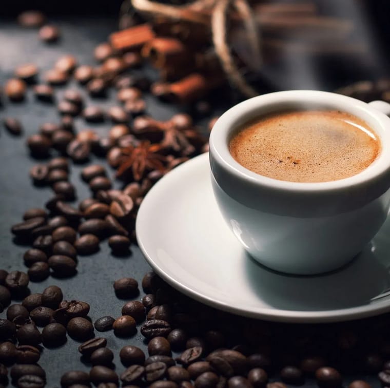 Tách sứ trắng đựng cà phê nóng có lớp crema, đặt trên đĩa lót giữa những hạt cà phê rang.