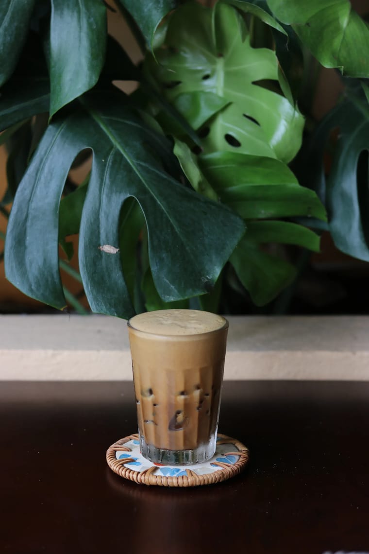 Ly cà phê đá với lớp bọt kem, đặt trên đế lót trước tán lá monstera.