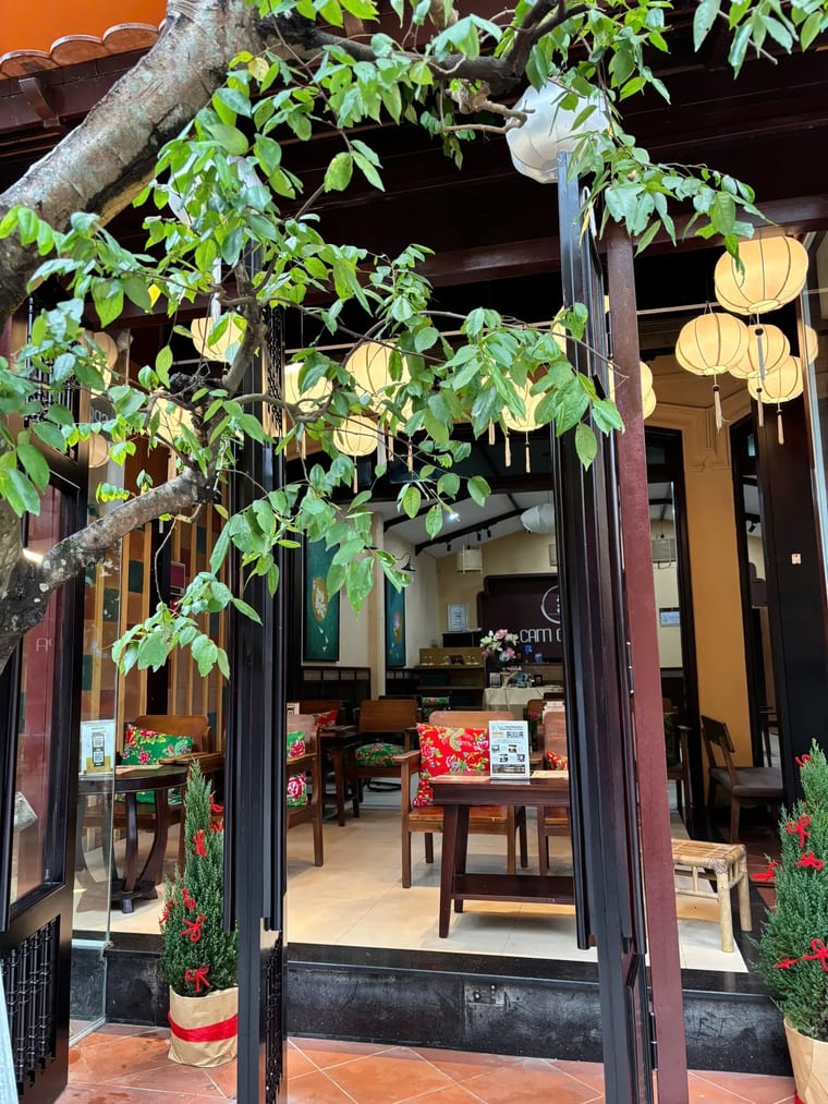 Lối vào ấm cúng của Cam On Cafe với ghế gỗ, nệm hoa và đèn lồng giấy toả sáng.