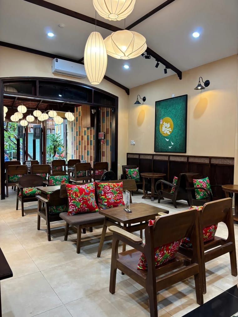 Khu vực ngồi trong nhà ấm cúng tại Cam On Cafe với ghế gỗ và nệm hoa nhiều màu.