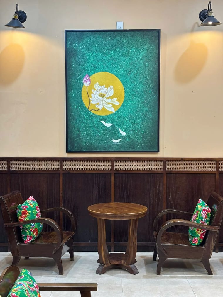 Góc ngồi trong nhà ấm cúng với ghế gỗ, bàn nhỏ và bức tranh sen lớn trên tường tại Cam On Cafe.