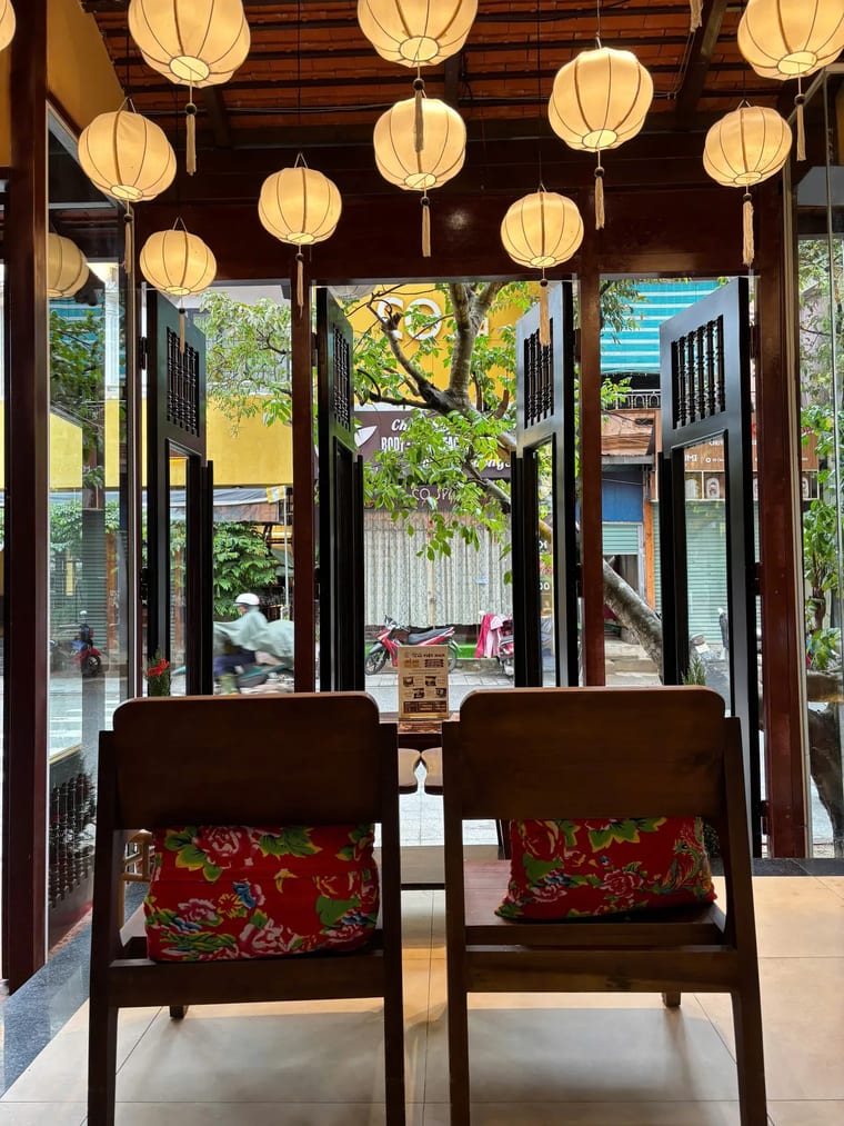 Hai chiếc ghế gỗ với nệm hoa truyền thống đặt dưới những chiếc đèn lồng trắng trong Cam On Cafe.