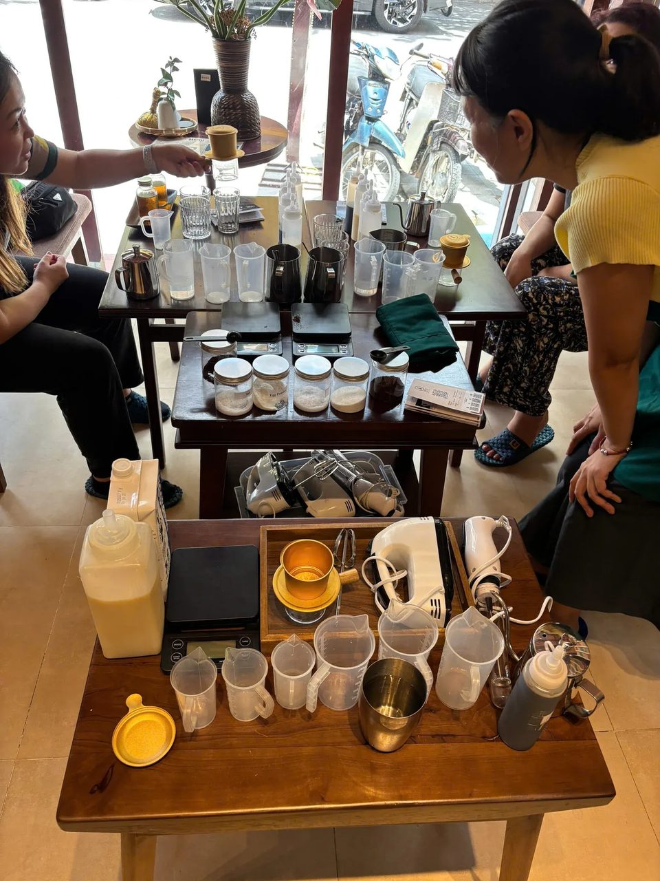 Học viên quây quanh những chiếc bàn gỗ đã bày sẵn phin, cân và cốc đong cho buổi học pha cà phê tại Cam On Cafe.